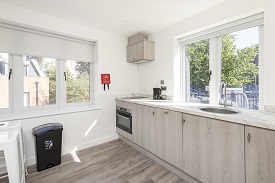 A kitchen in the Old Vicarage. The kitchen unit is against the wall with windows. The kitchen has an oven/hob, extractor fan, sink, kettle and cupboard space. The room also has a stool and bin.